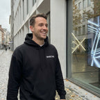 Lächelnder Mann trägt den schwarzen UN4oRGETTABLE Heavy Hoodie für AI Rebels in urbanem Streetwear-Look.